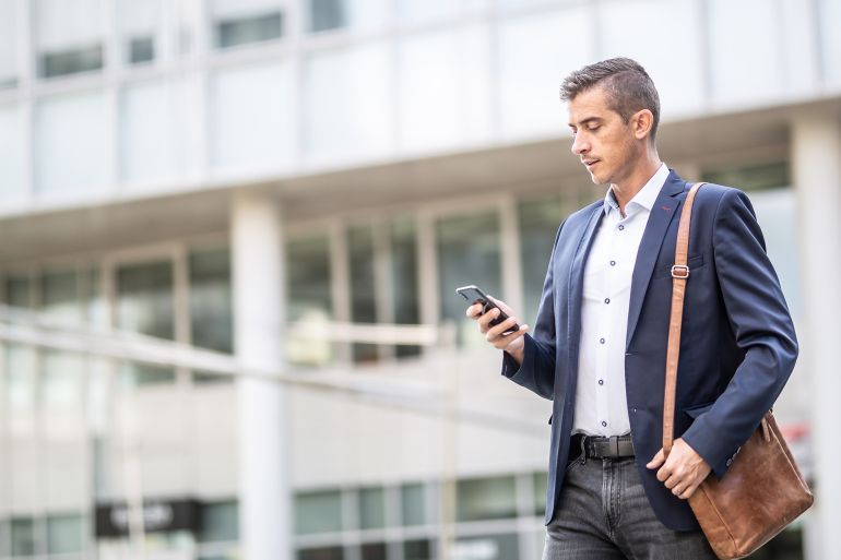Man in casual business attire checks his phone walking outdoors in an office buildings environment.; Shutterstock ID 2034828833; purchase_order: aj; job: ; client: ; other: