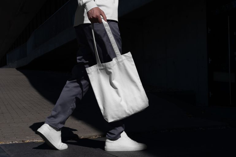 Man walking with a blank tote bag mockup template in an urban outdoor environment with sharp sunlit shadows. Isolated surface to place your design. ; Shutterstock ID 2533872383; purchase_order: aj; job: ; client: ; other: