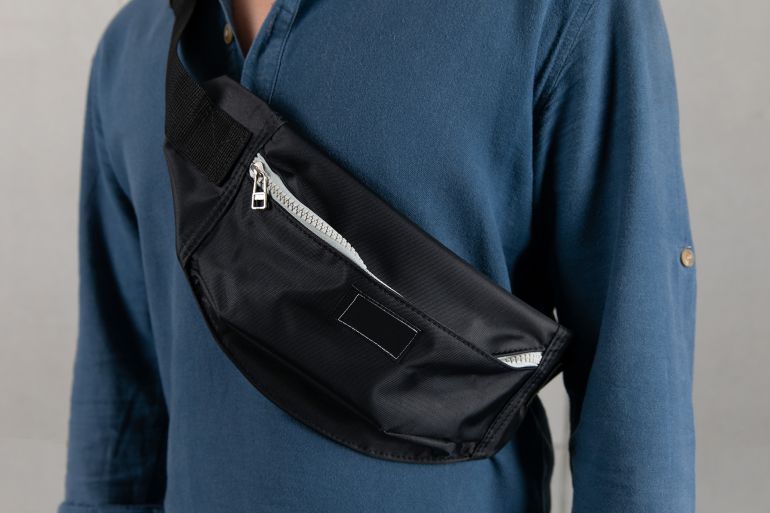 Closeup studio shot of male model in blue long sleeve shirt hanging trendy urban small black crossbody strap casual fanny pack bag in front gray background.; Shutterstock ID 2024715089; purchase_order: aj; job: ; client: ; other: