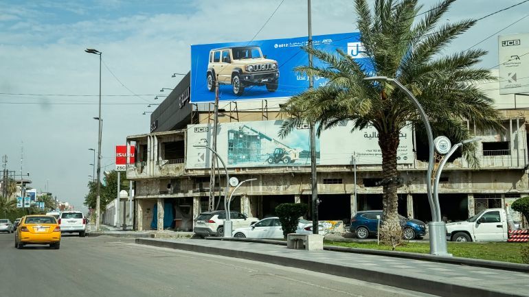 Baghdad, Iraq - March 20, 2025: Busy street in Baghdad showcases commercial properties, parked cars, palm trees, and vibrant advertisements on buildings.