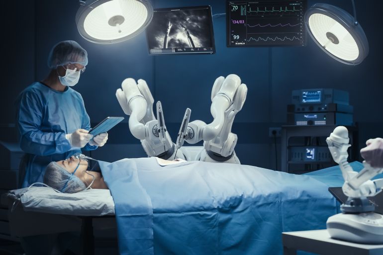 Surgeon Wearing AR Headset, Using High-Precision Remote Controlled Robot Arms To Operate On Patient, While His Colleague Checking Vitals On Tablet In Hospital. Doctors Working With Robotic Limbs.