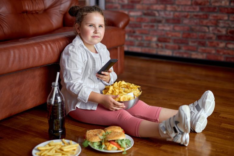 Fat girl eating chips from bowl while sitting on floor in living room, side view. - - سمنة الأطفال Overweight caucasian child in casual clothes enjoy leading unhealthy lifestyle, eat junk food and watch tv.