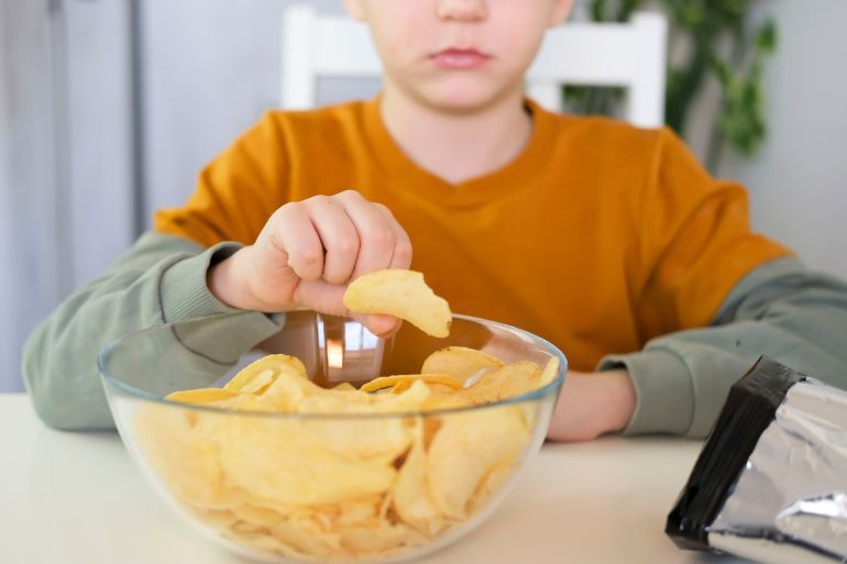 - سمنة الأطفال Close-up of hungry child putting potato chips into his mouth while sitting at table. Boy happily eats fast food, junk food at home. Childhood obesity concept. Selective focus.